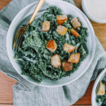 Kale Caesar Salad with fresh baked croutons in a white bowl on a wood table with a kitchen towel. Kale Caesar salad recipe by Farmer's Market Society. Market Inspired Meals.