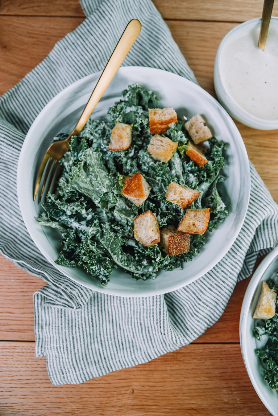 Kale Caesar Salad with fresh baked croutons in a white bowl on a wood table with a kitchen towel. Kale Caesar salad recipe by Farmer's Market Society.  Market Inspired Meals.